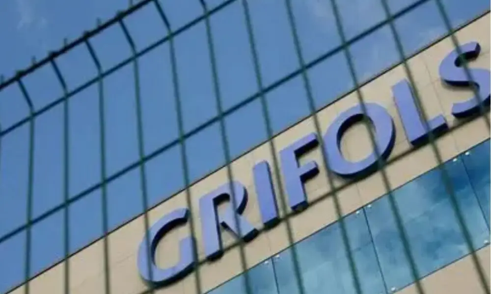 Close-up of a modern glass building exterior with large blue letters on the facade, viewed through a wire fence.