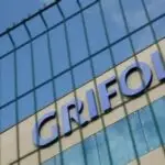 Close-up of a modern glass building exterior with large blue letters on the facade, viewed through a wire fence.