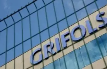 Close-up of a modern glass building exterior with large blue letters on the facade, viewed through a wire fence.
