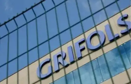 Close-up of a modern glass building exterior with large blue letters on the facade, viewed through a wire fence.