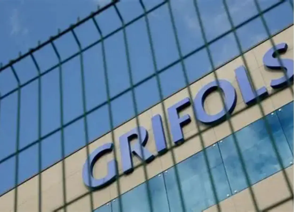 Close-up of a modern glass building exterior with large blue letters on the facade, viewed through a wire fence.