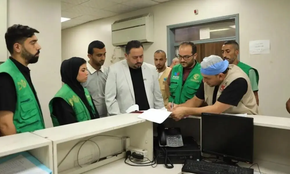 Group of aid workers in green vests gather around a reception desk as a man signs papers.