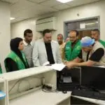 Group of aid workers in green vests gather around a reception desk as a man signs papers.