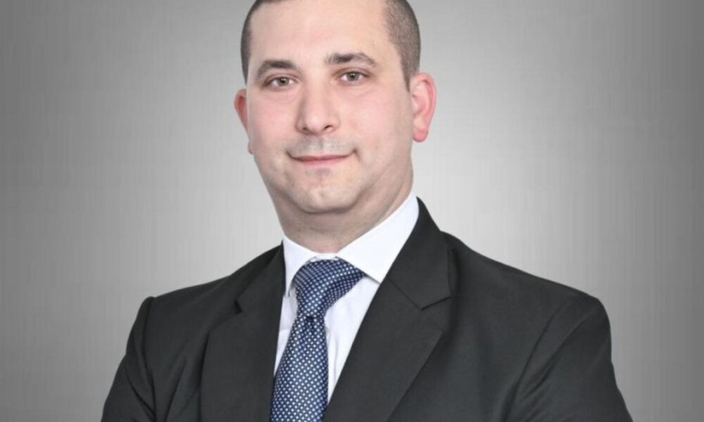 Professional headshot: man in a dark suit and blue tie against a gray gradient background, looking at camera with a slight smile.