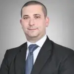 Image Sources - GlobeNewsWire.com - Base Markets Appoints Nazim Moussaoui as Head of Premium Clients and Partnerships - MENAWireNOW.com - MENAWireNOW Professional headshot: man in a dark suit and blue tie against a gray gradient background, looking at camera with a slight smile.