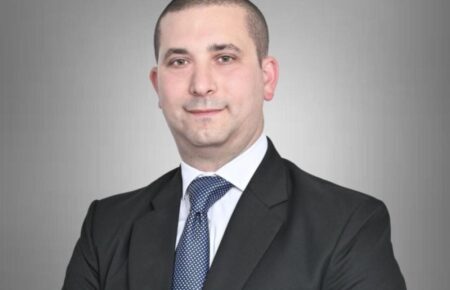 Professional headshot: man in a dark suit and blue tie against a gray gradient background, looking at camera with a slight smile.