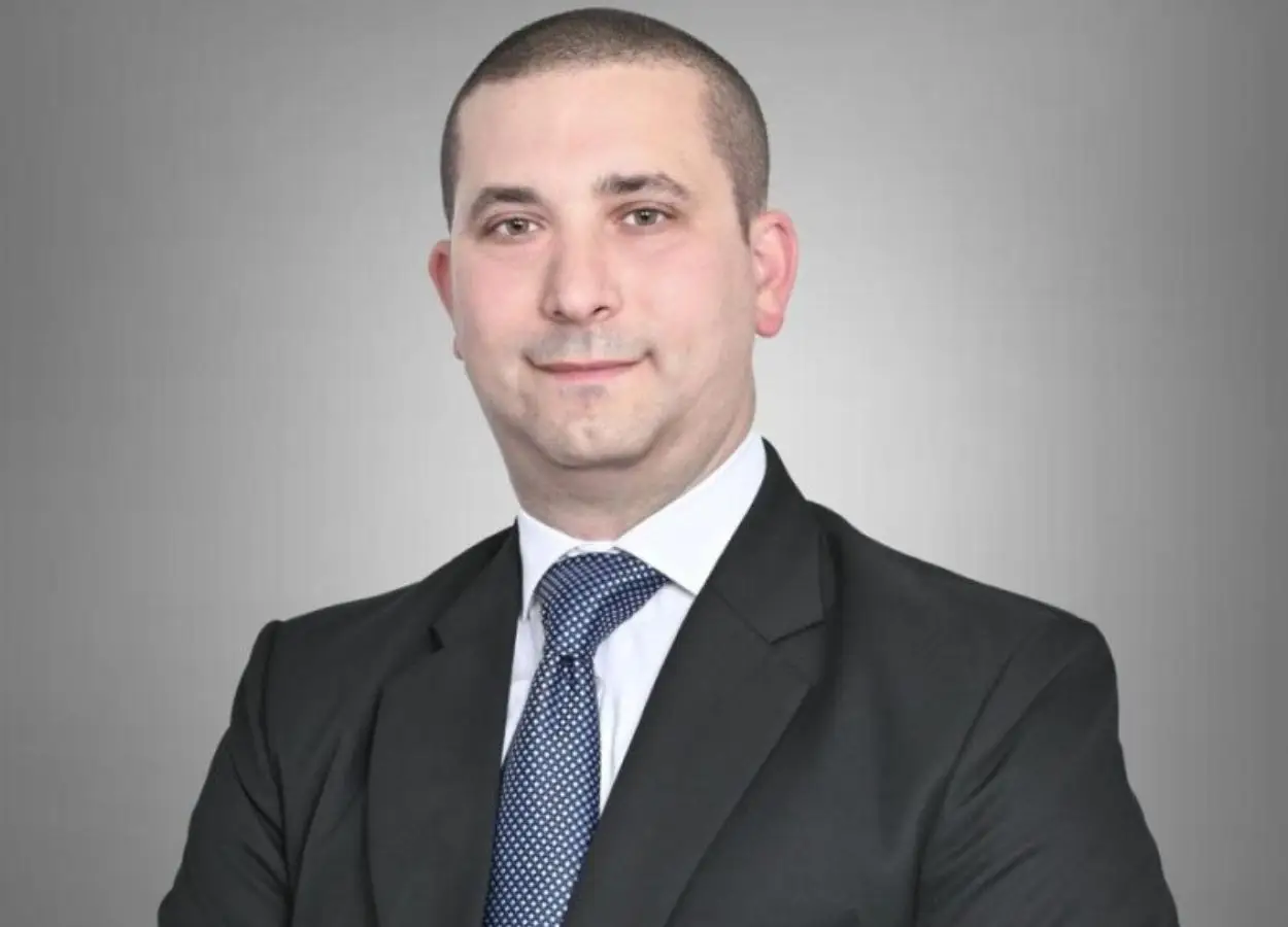 Professional headshot: man in a dark suit and blue tie against a gray gradient background, looking at camera with a slight smile.