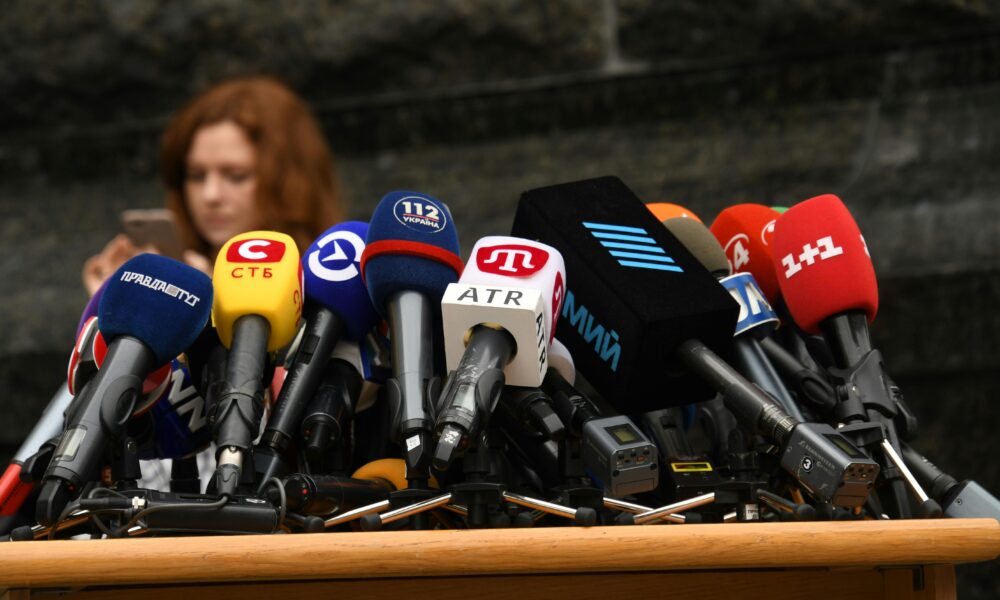 Dense cluster of colorful microphones on a wooden podium, ready for a press conference.