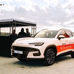 White SUV with orange SouvEast branding parked by a riverside event stage, people chatting in the background