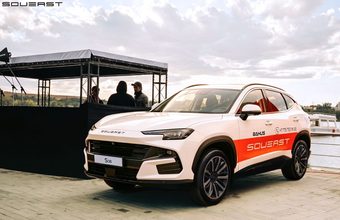 سيارة دفع رباعي بيضاء تحمل شعار SouvEast البرتقالي متوقفة بجوار منصة فعاليات على ضفة النهر، والناس يتحدثون في الخلفية