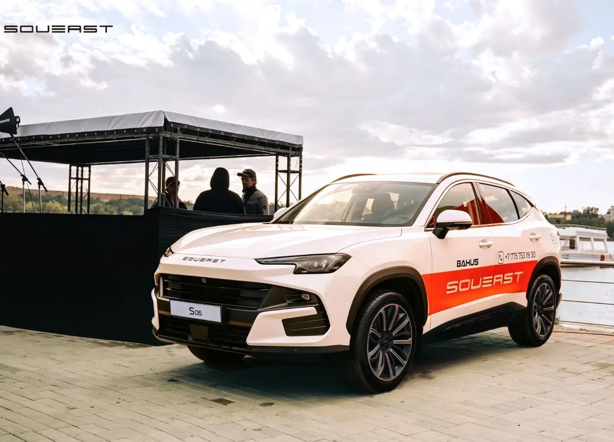 White SUV with orange SouvEast branding parked by a riverside event stage, people chatting in the background