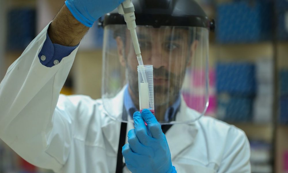 Scientist in a white lab coat with a face shield and blue gloves uses a pipette to transfer liquid into a graduated cylinder in a laboratory.