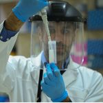 Scientist in a white lab coat with a face shield and blue gloves uses a pipette to transfer liquid into a graduated cylinder in a laboratory.