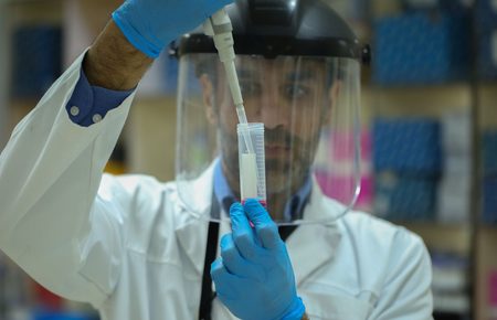 Scientist in a white lab coat with a face shield and blue gloves uses a pipette to transfer liquid into a graduated cylinder in a laboratory.
