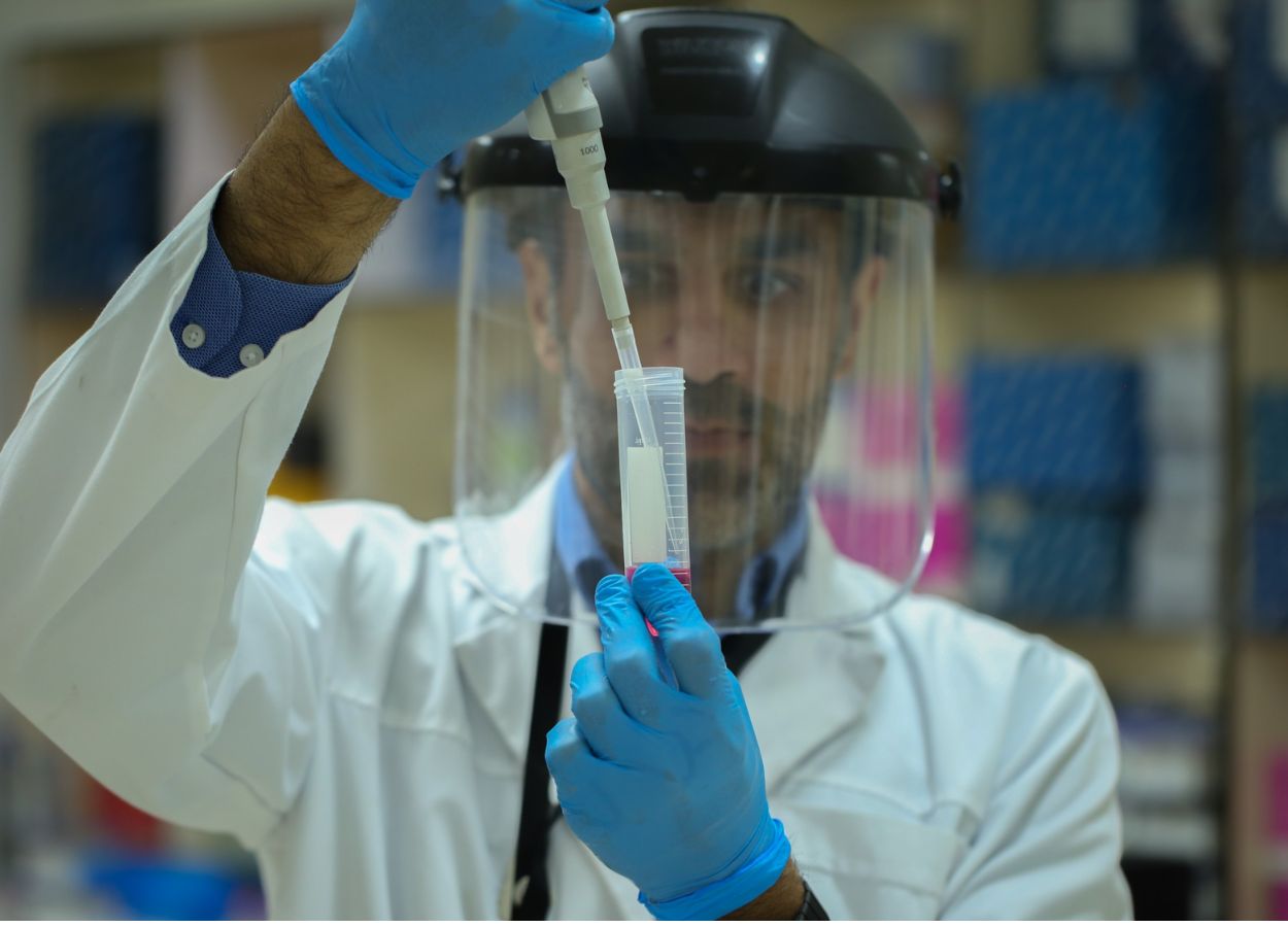 Scientist in a white lab coat with a face shield and blue gloves uses a pipette to transfer liquid into a graduated cylinder in a laboratory.