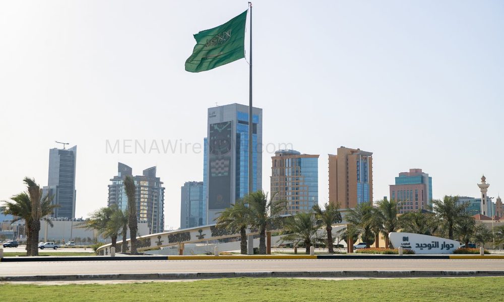 Green Saudi Arabia flag waving on a tall flagpole above a landscaped roundabout with palm trees and a cluster of modern high-rise buildings in the background