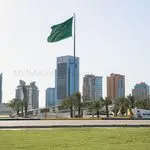 Green Saudi Arabia flag waving on a tall flagpole above a landscaped roundabout with palm trees and a cluster of modern high-rise buildings in the background