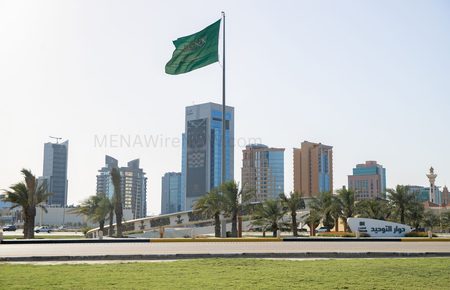 Green Saudi Arabia flag waving on a tall flagpole above a landscaped roundabout with palm trees and a cluster of modern high-rise buildings in the background