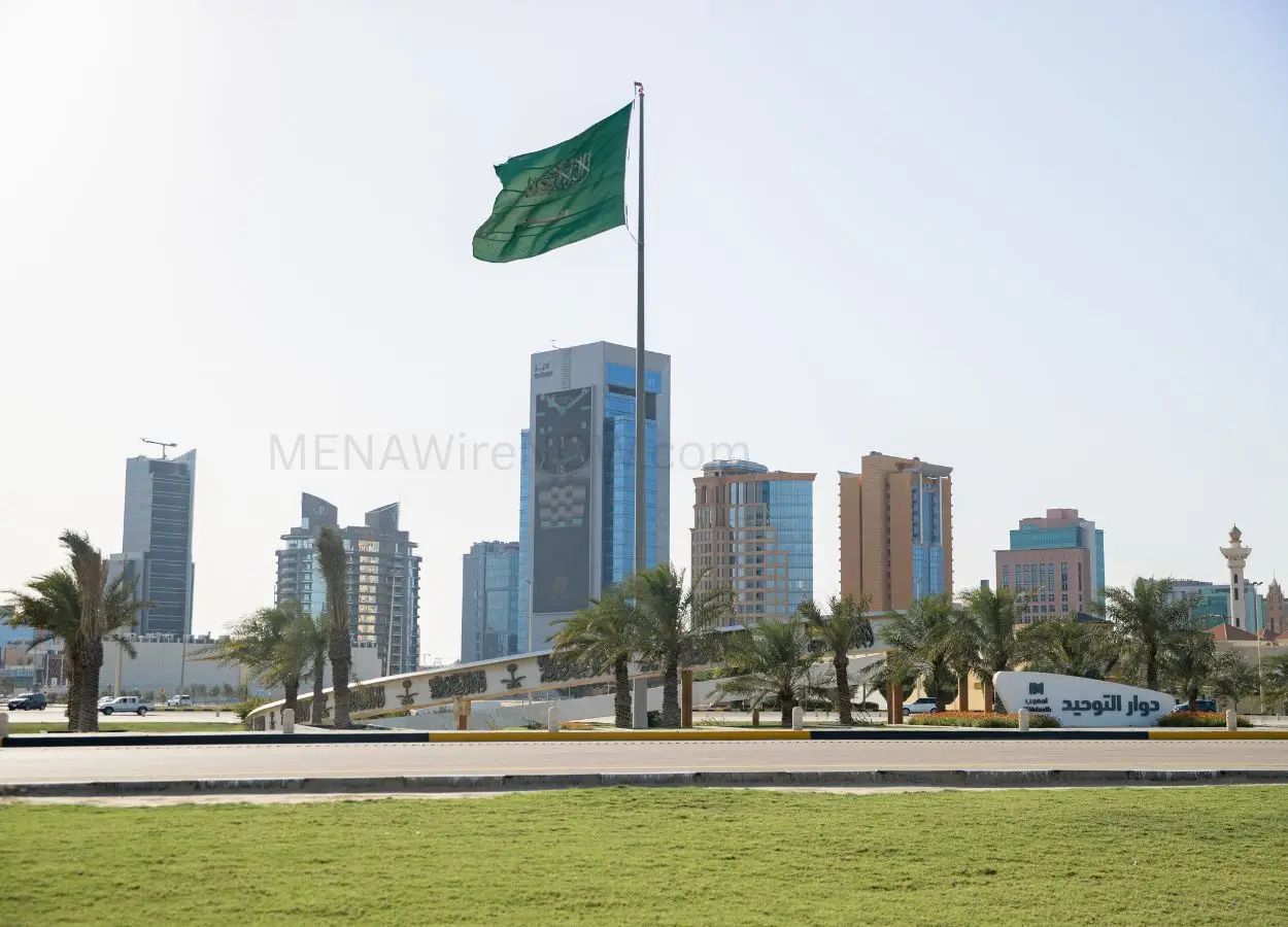 Green Saudi Arabia flag waving on a tall flagpole above a landscaped roundabout with palm trees and a cluster of modern high-rise buildings in the background