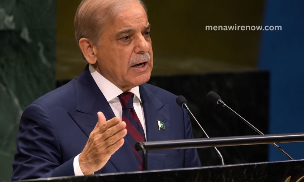 Man in a navy suit speaks at a podium with two microphones, gesturing with his hand; small Pakistan flag pin on his lapel.