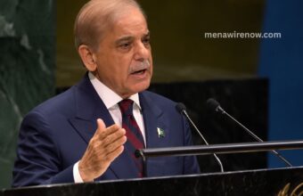 Man in a navy suit speaks at a podium with two microphones, gesturing with his hand; small Pakistan flag pin on his lapel.