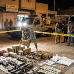 Night scene of a police raid: seized weapons and packages laid out on cloth, officers in tactical gear securing the area behind yellow caution tape.